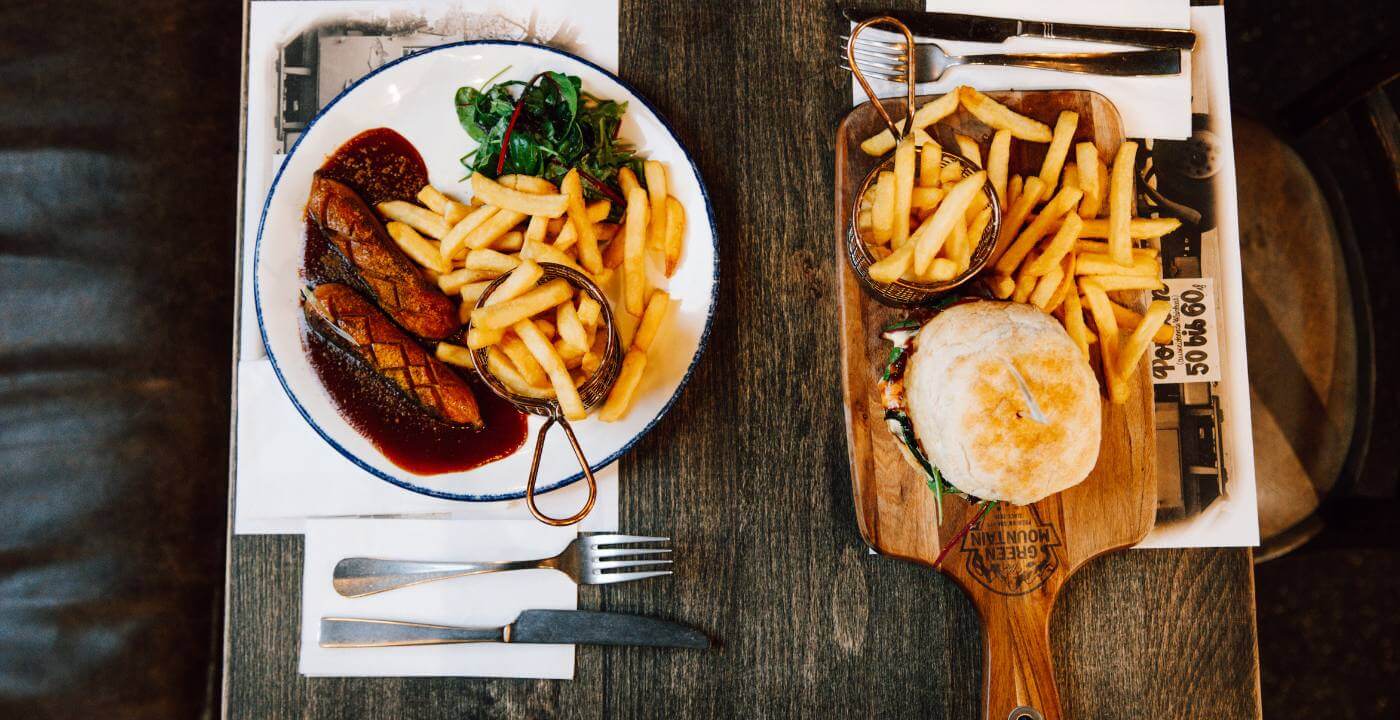 Ein veganer Burger mit Pommes auf einem Holzbrett und eine Bratwurst mit Pommes auf einem Teller